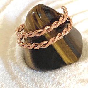 May include: A copper ring featuring a double-twisted wire design. The band is constructed from two strands of metallic copper wire woven together in a continuous rope-like pattern, creating a textured surface with a warm, reddish-brown finish.