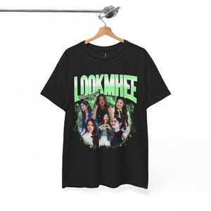 May include: Black t-shirt with a green and white graphic that reads "LOOKMHEE" and a collage of images. The design has a vintage, distressed appearance. The t-shirt is suspended from a wooden hanger.