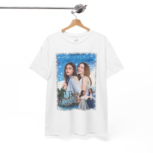 May include: White t-shirt with a graphic print of two women and text within a blue frame. The graphic includes the words "4 ELEMENTS" and "True Waters". The t-shirt is suspended on a wooden hanger.