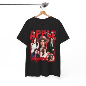 May include: Black t-shirt with a red graphic design featuring the word "APPLE" and images of a person. The design includes the name "Din" and "Lapisara". The t-shirt is displayed on a wooden hanger.