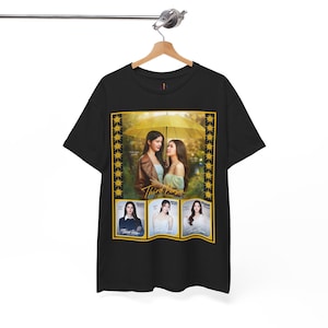 Op de afbeelding: Een zwart t-shirt met korte mouwen en een grafische print van twee personen onder een gele paraplu met de tekst Third Person. Daaronder staan drie ingelijste portretten van personen genaamd Gam, Mook en Piglet met de tekst Third Person.