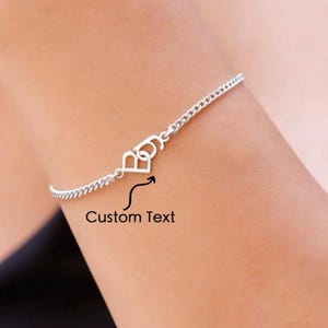 Op de afbeelding: Een zilveren kettingarmband met de letters "B" en "O" verbonden door een hartvormig ontwerp. De tekst "Custom Text" staat onder de armband.