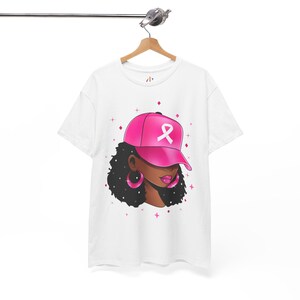 Puede incluir: Camiseta blanca con una ilustración rosa y blanca de una mujer con una gorra de béisbol rosa con una cinta rosa. La mujer tiene el pelo castaño oscuro y grandes pendientes de aro.