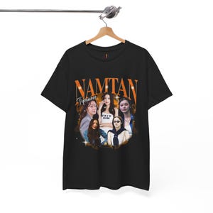 Könnte beinhalten: Schwarzes T-Shirt mit einem orangefarbenen Aufdruck des Wortes "NAMTAN" und dem Namen "Tipnaree" darunter. Der Aufdruck enthält Bilder von mehreren Frauen. Das T-Shirt hängt an einem Holzkleiderbügel.