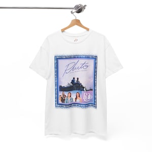 Può includere: T-shirt bianca con un design grafico con la parola "Pluto" in un font stilizzato. Il design include immagini di persone e testo in lingua straniera, il tutto all'interno di una cornice blu. La t-shirt è appesa a una gruccia di legno.