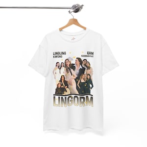 Puede incluir: Camiseta blanca con una foto de un grupo de mujeres. La camiseta tiene el texto "LINGLING KWONG" y "ORM KORNNAPHAT" en la parte superior, con la palabra "LINGORM" en letras doradas en la parte inferior. La camiseta está colgada en una percha de madera.