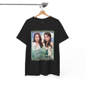 May include: Black t-shirt with a movie poster design featuring two women and text in a foreign language. The poster has a green and glittery border. The t-shirt is suspended from a wooden hanger.