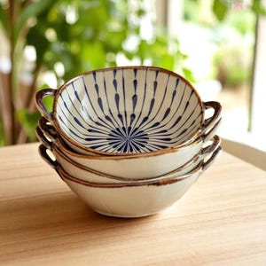 Può includere: Una pila di quattro ciotole in ceramica bianco sporco con manici. Ogni ciotola presenta un motivo floreale blu al centro e un bordo marrone. Le ciotole sono impilate su una superficie di legno.