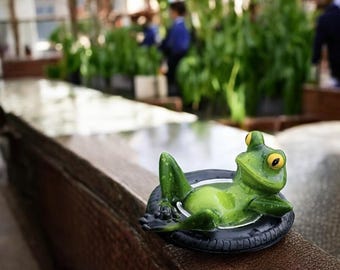 Könnte beinhalten: Eine grüne Froschfigur, die sich in einem schwarzen Reifeninneren entspannt. Der Frosch hat gelbe Augen und befindet sich auf einer Holzoberfläche. Der Hintergrund ist verschwommen, was auf eine Umgebung im Freien hindeutet.