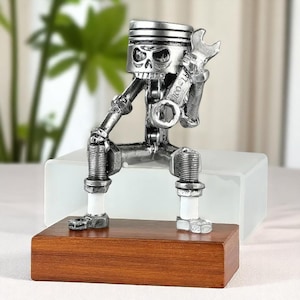 以下が含まれることがあります： エンジン部品で作られた、レンチを持った骸骨の銀色の金属彫刻。 頭蓋骨の形をした頭はピストンで、脚はスパークプラグです。 彫刻は木製の台座に置かれています。