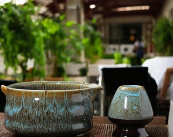 May include: A ceramic matcha bowl and a matching chawan, both with a blue and brown glaze. The bowl has a spout and handles. The chawan has a rounded shape and a dark brown base. The set is on a brown surface.