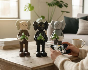 May include: Three KAWS Companion figures in brown, black, and gray, each holding a green bundle. The figures have X-shaped eyes and are displayed on a wooden table. A person is taking a photo of the figures with a smartphone.