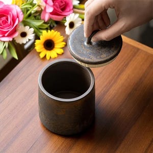 Pode incluir: Um pote de cerâmica marrom escuro com uma tampa levantada. O pote cilíndrico tem uma superfície texturizada. A tampa tem uma pequena alça. Flores rosas, amarelas e brancas ao fundo. O pote tem cerca de 10 cm de altura.