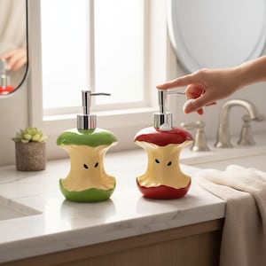 Könnte beinhalten: Zwei Keramik-Seifenspender in Form von Apfelkernen. Einer ist grün und gelb, der andere rot und gelb. Beide haben silberne Pumpköpfe und schwarze Kerndetails. Auf einer weißen Arbeitsplatte.