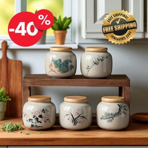 Puede incluir: Cinco tarros de cerámica con tapas de madera, cada uno adornado con un diseño floral o botánico único en tonos verdes, rosas y grises. Los tarros se exhiben en una estantería y encimera de madera.