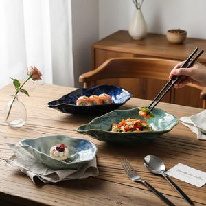 Può includere: Tre piatti di ceramica a forma di foglia nei toni del blu e del verde sono esposti su un tavolo di legno. Un piatto contiene rotoli di sushi, un altro ha un dessert e il terzo un piatto di verdure. Le bacchette prendono il cibo.