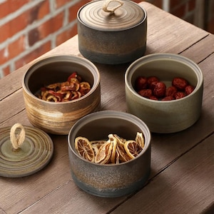 以下が含まれることがあります： 蓋付きの陶器製の保存瓶のコレクションで、乾燥フルーツが入っています。瓶は茶色とグレーの様々な色合いで、素朴で質感のある仕上がりです。1つの蓋にはロープの取っ手が付いています。木製の表面に配置されています。
