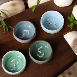May include: Four ceramic bowls in shades of blue and green, each with a painted koi fish design. The bowls are arranged on a dark wooden surface, with green leaves and white objects in the background. The bowls have a smooth, glossy finish.