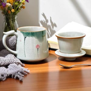 Puede incluir: Un juego de té de cerámica con una taza con un asa única y un infusor de té. La taza tiene un esmalte craquelado blanco con detalles florales verdes y rosas. Una cuchara pequeña descansa sobre la superficie de madera.