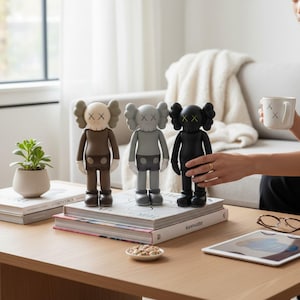 May include: Three KAWS Companion figures in brown, grey, and black, each with X-shaped eyes and a rounded head. They stand on a stack of books on a wooden table. A white mug with X markings is held in the background.