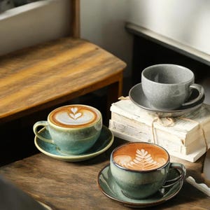 May include: Three ceramic coffee cups and saucers in green and grey hues, filled with coffee and latte art. The cups are on a wooden surface, with a stack of books in the background. The latte art features leaf and heart designs.
