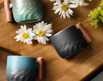 May include: Three ceramic mugs with wooden handles in shades of blue, black, and green. The mugs have a textured wave design and are arranged on a wooden tray with white daisies.