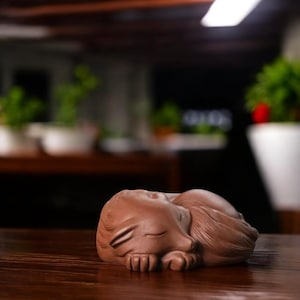 May include: A brown ceramic sculpture of a sleeping cat, curled up on a wooden surface. The cat's eyes are closed, and its paws are tucked beneath its body. The sculpture is detailed and realistic, with a smooth finish.