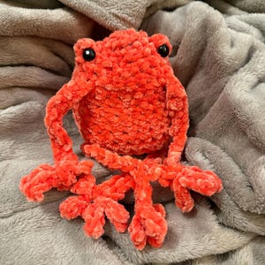 May include: A plush, coral-colored frog with black eyes. The handmade frog has outstretched arms and legs, and is made of a soft, textured material. The frog is sitting on a gray, fuzzy blanket.