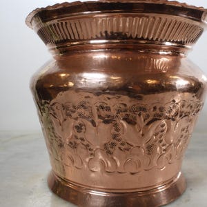May include: A copper-colored decorative vase with a wide, fluted rim and a bulbous body. The vase features an embossed floral design around the middle and a rounded base. The copper has a polished, reflective surface.