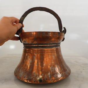 Op de afbeelding: Een verweerde koperen emmer met een brede, uitlopende basis en een donker, getextureerd ijzeren handvat. Het metalen oppervlak vertoont een warme, oranje patina met zichtbare tekenen van slijtage en oxidatie. Gegraveerde tekst is zichtbaar onderaan.