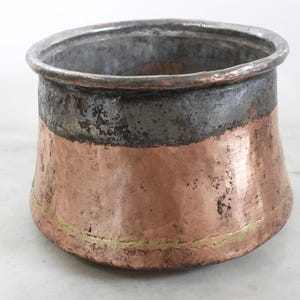 May include: A hand-hammered copper pot with a wide, open top and a rounded base. The upper rim features a dark, weathered metallic band, while the lower body displays a warm copper tone with a thin, horizontal brass-coloured accent line near the bottom.