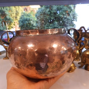 Op de afbeelding: Een gehamerde koperen pot met twee kleine handvatten. De pot heeft een afgeronde vorm en een donkere, verouderde patina. Andere decoratieve items van messing zijn zichtbaar op de achtergrond.