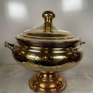 Op de afbeelding: Een goudkleurige, gehamerde metalen serveerschaal met een deksel en een voet. De kom heeft twee kleine handvatten en een afgeronde knop op het deksel. Het oppervlak van de kom heeft een getextureerd, kuiltjesachtig uiterlijk.