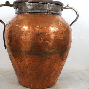 May include: A large, antique copper pot with a hammered texture. The pot has a silver-colored rim and lid, and two curved handles. The copper has a rich, warm color with some green and blue oxidation.
