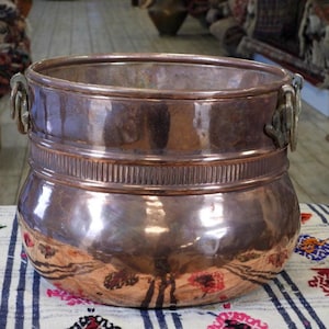 May include: A round, polished copper pot featuring a wide, open top and a rounded body. The vessel includes a horizontal band with vertical ribbed detailing around the upper section and two metal ring handles attached to the sides of the rim.