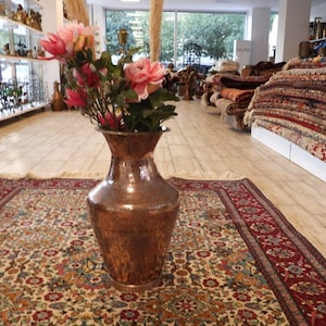 Peut inclure: Un grand vase en cuivre rempli de fleurs roses et rouges est posé sur un tapis à motifs dans une boutique. Le tapis a un fond crème avec des motifs floraux et une bordure rouge. Des tapis sont empilés en arrière-plan.