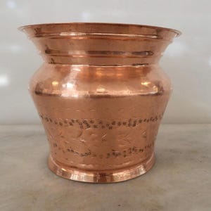 May include: A polished copper pot with a wide, flared rim and a rounded body. The exterior surface features a horizontal band of intricate, etched leaf patterns and cross-hatched detailing, set against a smooth, reflective metallic finish.