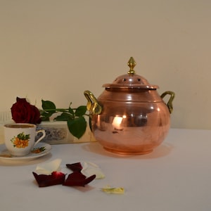 Peut inclure: Une marmite en cuivre avec un couvercle et des poignées en laiton, à côté d'une tasse et d'une soucoupe à motifs floraux. Une rose rouge et un livre sont également dans l'image. La marmite a un bouton décoratif sur le dessus.