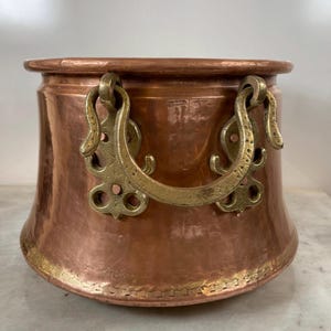 Op de afbeelding: Een ronde koperen pot met een brede, uitlopende rand en een gehamerde textuur. De pot is voorzien van twee sierlijke, gebogen messing handgrepen die aan de zijkanten zijn bevestigd met decoratieve platen en kleine koperen klinknagels.