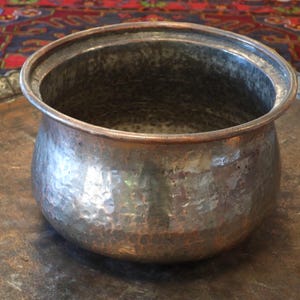 May include: A round, hammered metal pot with a wide, open top and a slightly flared rim. The exterior surface features a textured, dimpled finish with a metallic silver and copper-toned appearance, showing signs of age and natural oxidation.