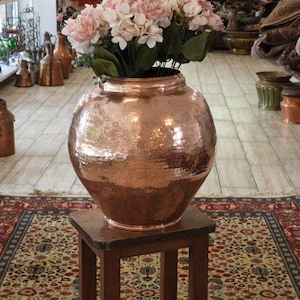 May include: A large, hammered copper vase filled with pink flowers sits on a small wooden stool. The vase has a rounded shape and a wide opening. The background includes a patterned rug and other copper decorative items.