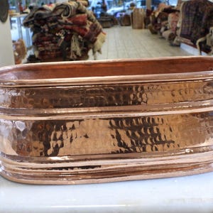 May include: A hammered copper planter, rectangular with rounded edges. It has a polished finish and black handles. The copper is a warm, reddish-brown colour. The planter is likely for decorative use. The length is approximately 60 cm.