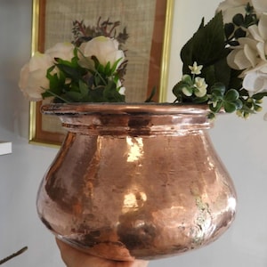 May include: A round, hammered copper pot with a wide, flared rim. The metallic surface reflects light, showing a textured, uneven finish. Inside the pot, there are several white artificial flowers with green leaves and stems extending upwards.
