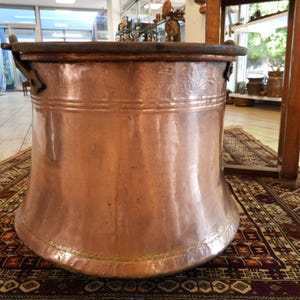 May include: A large copper cauldron with a wide base and a dark rim. The cauldron has a polished, reflective surface and is displayed on a patterned rug. The copper pot is a vintage kitchenware item.