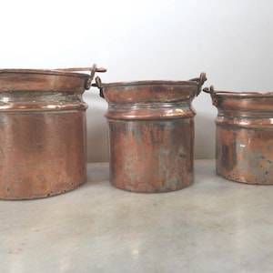Op de afbeelding: Een set van drie koperen emmers in oplopende grootte met een gepolijste metalen afwerking. Elke emmer heeft een cilindrisch lichaam met een duidelijke horizontale rand nabij de bovenkant en twee kleine metalen handvatten aan de zijkant.