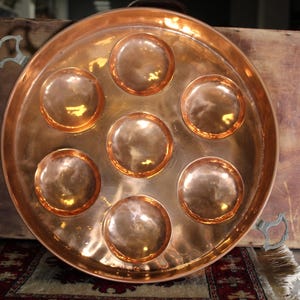 May include: A large, round copper tray with seven circular indentations. The tray has a polished, reflective surface and sits on a wooden surface. The tray is likely used for serving food or displaying items.