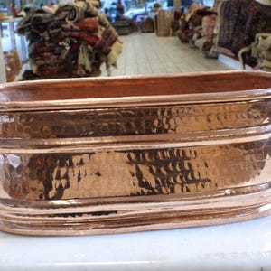 May include: An oval-shaped copper planter or basin featuring a hammered texture on its exterior surface. The vessel includes two dark metal handles attached to the sides and horizontal ridges running along the body, providing a metallic finish.