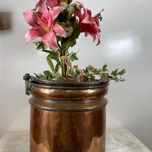 May include: A copper-coloured metal container with a dark rim, filled with pink lilies and greenery. The container is cylindrical with a flared top and a handle. The lilies are a vibrant pink, and the greenery adds a fresh touch. The container is approximately 25 cm tall.
