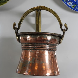 Peut inclure: Un seau en cuivre martelé avec une base évasée et un bord large. Il est doté d'une anse pivotante en laiton fixée à un support mural vertical en laiton. La poignée porte l'inscription gravée L'NOEL ainsi que d'autres marques.