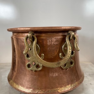 Op de afbeelding: Een koperen pot met messing handvatten. De pot heeft een afgeronde body met een uitlopende basis en een gerolde rand. De messing handvatten zijn versierd en met klinknagels bevestigd. De pot is waarschijnlijk om te koken.